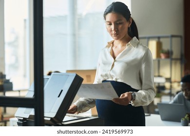 Young pregnant Asian businesswoman in smart casualwear looking at financial document while standing by xerox machine in office - Powered by Shutterstock