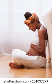 Young pregnant black woman seated on the floor touching her belly- African people
