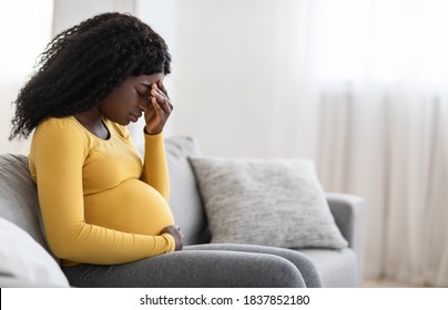 Young pregnant black woman suffering from headache or migraine, feeling sick, sitting on sofa at home, free space. Sad african american expecting lady touching her forehead and big tummy, home alone