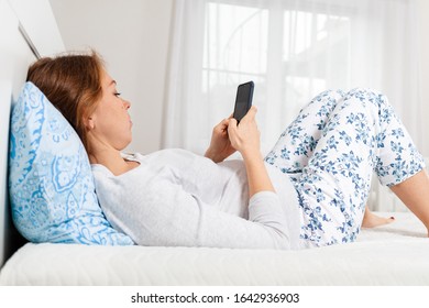 A young pregnant Caucasian woman is lying on a bed and using her smartphone. A white room and a window in the background. The concept of the Internet and social networks. Close up - Powered by Shutterstock