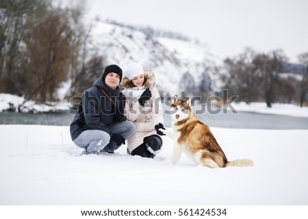A young pregnant couple walking in the woods with the dog red Husky with blue eyes. Snowy morning in the forest. A girl and a guy dressed in warm clothes