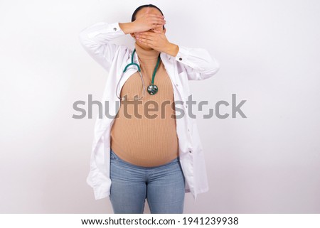 young pregnant doctor woman wearing medical uniform against white background Covering eyes and mouth with hands, surprised and shocked. Hiding emotions.