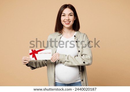 Young pregnant expectant woman future mom wear grey shirt with belly stomach tummy with baby hold store gift coupon voucher card isolated on plain beige background. Maternity family pregnancy concept