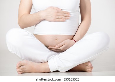 Young pregnant fitness model in sportswear doing yoga or pilates training, sitting in Easy Pose, Sukhasana, touching her belly, breathing, gray background, studio shot, close-up