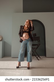 young pregnant girl in black jacket and jeans stands casual on a mint wall background with arch and director's chair at sunny day and looks apart with hand near hair. pregnant concept, free space