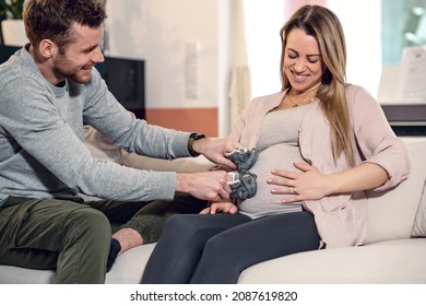 Young pregnant happy couple sitting on couch husband stroking baby belly of blond laughing wife with baby shoes
