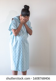 A young pregnant mixed race African American woman wearing a hospital gown or robe puts or hides her face in her open palms as she hunches over crying tears of sadness or depression.