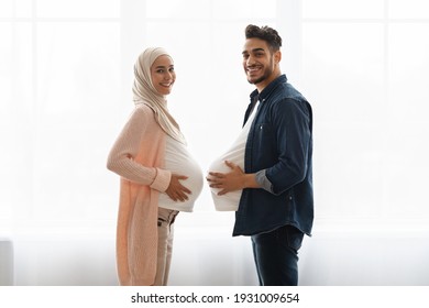 Young pregnant muslim woman and her husband having fun at home, comparing belly sizes. Cheerful arab man with big fake tummy standing next to his expecting wife near window, smiling at camera