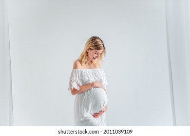 A Young Pregnant Woman In Anticipation Of A Baby In A White Dress, Hugs Belly. Fashion Girl In A Minimalistic, Isolated Interior Of A Cut Of Airy Fabrics. Easy, Happy Maternity Pregnancy, Surrogacy.