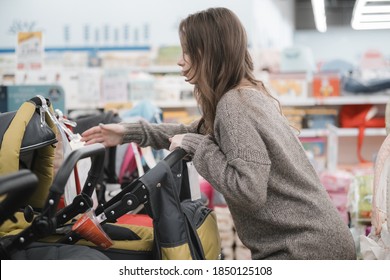 Young Pregnant Woman Carefully Choosing Infant Baby Stroller Or Pram Buggy For Newborn. Pregnancy And Shopping. Shopping For Expectant Mothers And Baby. Young Pregnant Woman Is Of Baby Store In Mall