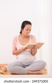 Young Pregnant Woman Chewing Gum And Reading A Book While Sitting On The Floor