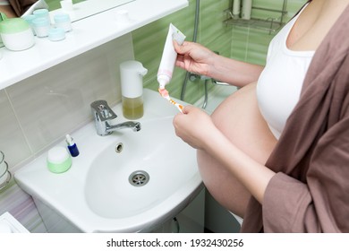 Young Pregnant Woman Dressed In A Robe In The Bathroom At Home. Cute Adult Girl In The Third Trimester Of Pregnancy In The Home Bathroom Brushing Her Teeth.