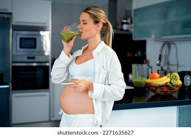 A young pregnant woman drinks a prepared vitamin smoothie and uses a smartphone standing in the kitchen at home. Healthy eating during pregnancy. - Powered by Shutterstock