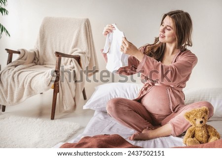 young pregnant woman with long hair in pajamas examines baby clothes for newborns while sitting on bed. mother expecting child. Collecting things for the maternity hospital. Preparing for childbirth