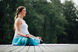 Young Pregnant Woman In The Lotus Position Is Practicing Yoga In The Forest Next To The River. Sitting On Yoga Mats On The Wooden Pier.