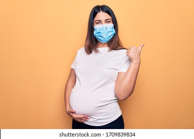 Young pregnant woman mother to be wearing protection mask for coronavirus disease pointing thumb up to the side smiling happy with open mouth