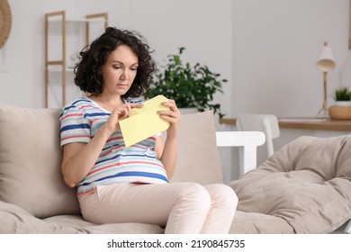 Young Pregnant Woman Opening Envelope On Couch At Home