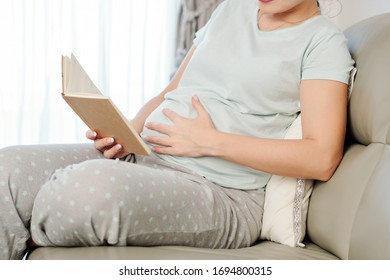 Young pregnant woman in pajamas touching her belly when reading a book or journal - Powered by Shutterstock