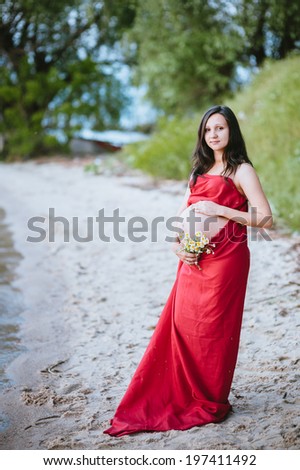 The young pregnant woman in red dress with a bouquet of wild daisies walks in the park by the lake. Riverside. Young pregnant woman walking on the beach