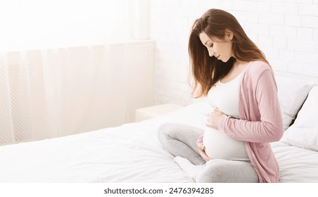 A young pregnant woman is seated on a bed in a bright, minimalist bedroom during the daytime. She is gently cradling her belly and appears deep in thought, copy space - Powered by Shutterstock