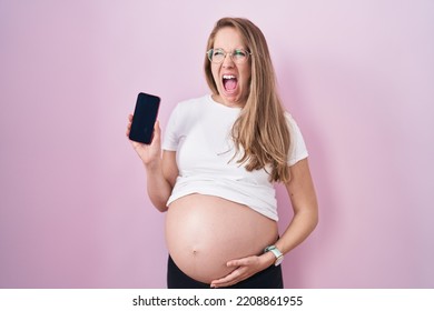 Young Pregnant Woman Showing Belly Holding Smartphone Angry And Mad Screaming Frustrated And Furious, Shouting With Anger. Rage And Aggressive Concept. 