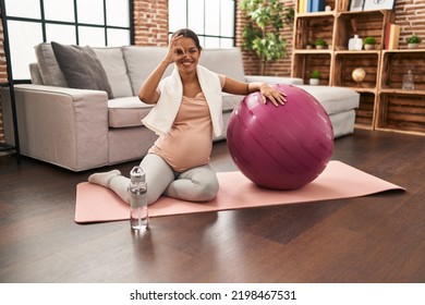 Young pregnant woman sitting on yoga mat with pilates ball smiling happy doing ok sign with hand on eye looking through fingers 