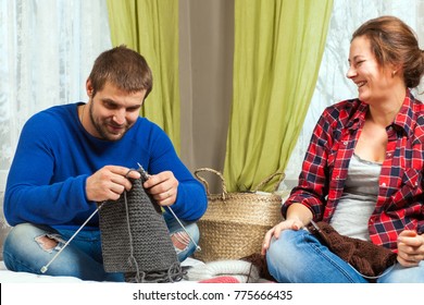 Young Pregnant Woman Smiles And Teaches Young Man In Blue Sweater And Jeans How To Knit With Knitting Needles From Natural Yarns Gray Sweater Sitting On Bed In Home Setting