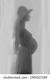 A Young Pregnant Woman Is Standing In Profile Behind A Transparent Curtain. The Image Is Blurry. No Face Visible. With A Wide-brimmed Hat. Monochrome. Black And White Photo. Studio.