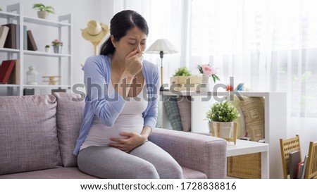 young pregnant woman suffering from toxicosis at home. asian japanese maternity lady having morning sickness covering mouth with hands while sitting on couch in living room. mom expect unborn baby