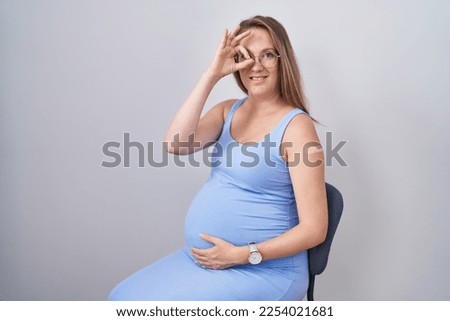 Young pregnant woman wearing band aid for vaccine injection smiling happy doing ok sign with hand on eye looking through fingers 