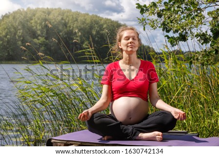 Young pregnant woman wearing sporty dress doing yoga exercise sitting in lotus position on a wooden pier at a forest lake meditating. Fitness and healthy lifestyle during pregnancy concept