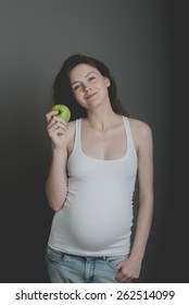 Young pregnant woman in white shirt and jeans on grey background holding green apple