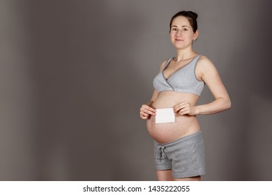 Young Pregnant Woman With A Wry Grin In A Modest Gray Suit Holding A White Sheet Of Paper That Looks Like An Ultrasound On A Dirty Gray Background