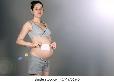 Young Pregnant Woman With A Wry Grin In A Modest Gray Suit Holding A White Sheet Of Paper That Looks Like An Ultrasound On A Dirty Gray Background