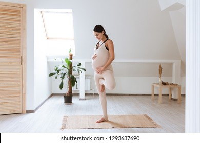 Young pregnant yoga woman working out at home doing prenatal Tree posture, Vrksasana yoga pose. Healthy Pregnancy concept. Pregnant Namaste gesture