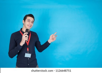 young presenter pointing at something while holding microphone and smiling to camera , on blue background