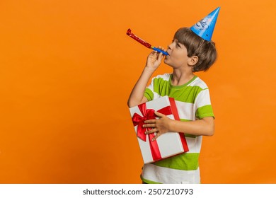 Young preteen child boy kid holding gift box wearing birthday cone hat blowing pipe celebrating success win victory. Excited lucky little children in anniversary party isolated on orange background