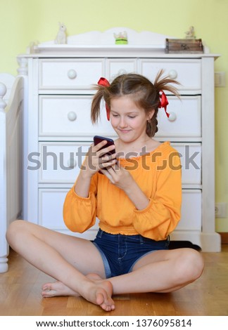 Young pre-teen girl in the bright orange  jacket with funny pigtails plaing with a mobile phone and making self-portrait and snapchat, sitting on the floor at the home