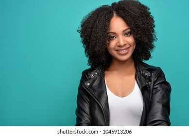 Young Pretty African American Millennial Woman With Natural Curly Hair Wearing Fashionable White Tank Top And Black Leather Jacket Smiling. Blue  Background.
