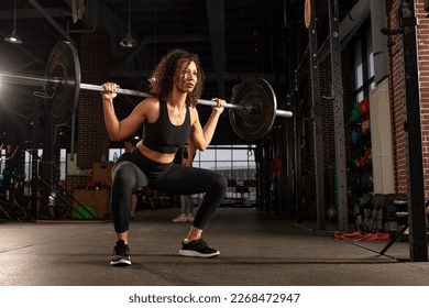 Young pretty african american sports woman in fitness gym lifting up empty bar in gym, doing warm up, barbell squat, butt workout - Powered by Shutterstock