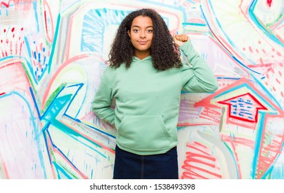 Young Pretty African American Woman Feeling Serious, Strong And Rebellious, Raising Fist Up, Protesting Or Fighting For Revolution Against Graffiti Wall