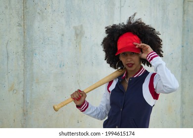 Young, pretty African American woman in baseball cap and jacket with baseball bat resting on shoulder against gray cement background.