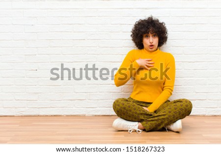 young pretty afro woman feeling shocked, astonished and surprised, with hand on chest and open mouth, saying who, me? sitting on the floor