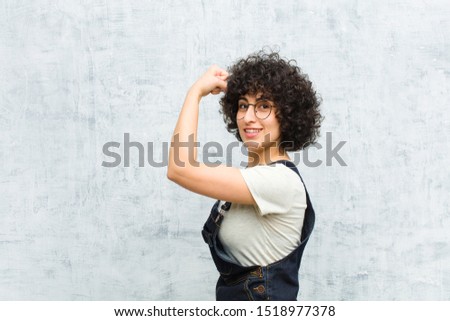 young pretty afro woman feeling happy, satisfied and powerful, flexing fit and muscular biceps, looking strong after the gym