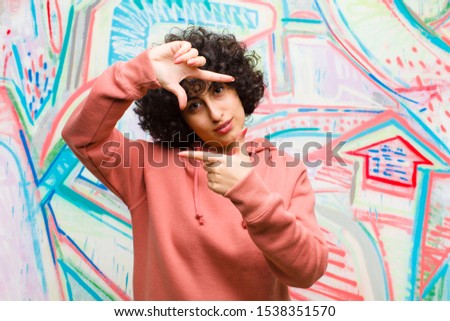 young pretty afro woman feeling happy, friendly and positive, smiling and making a portrait or photo frame with hands against graffiti wall