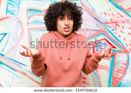 young pretty afro woman feeling clueless and confused, not sure which choice or option to pick, wondering against graffiti wall