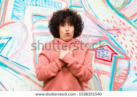 young pretty afro woman looking puzzled and confused, insecure and pointing in opposite directions with doubts against graffiti wall