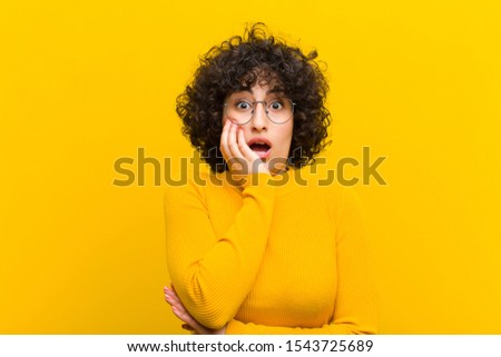 young pretty afro woman open-mouthed in shock and disbelief, with hand on cheek and arm crossed, feeling stupefied and amazed