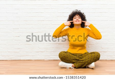 young pretty afro woman smiling confidently pointing to own broad smile, positive, relaxed, satisfied attitude sitting on the floor