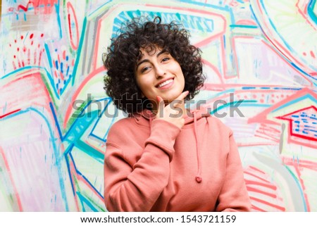 young pretty afro woman smiling, enjoying life, feeling happy, friendly, satisfied and carefree with hand on chin against graffiti wall